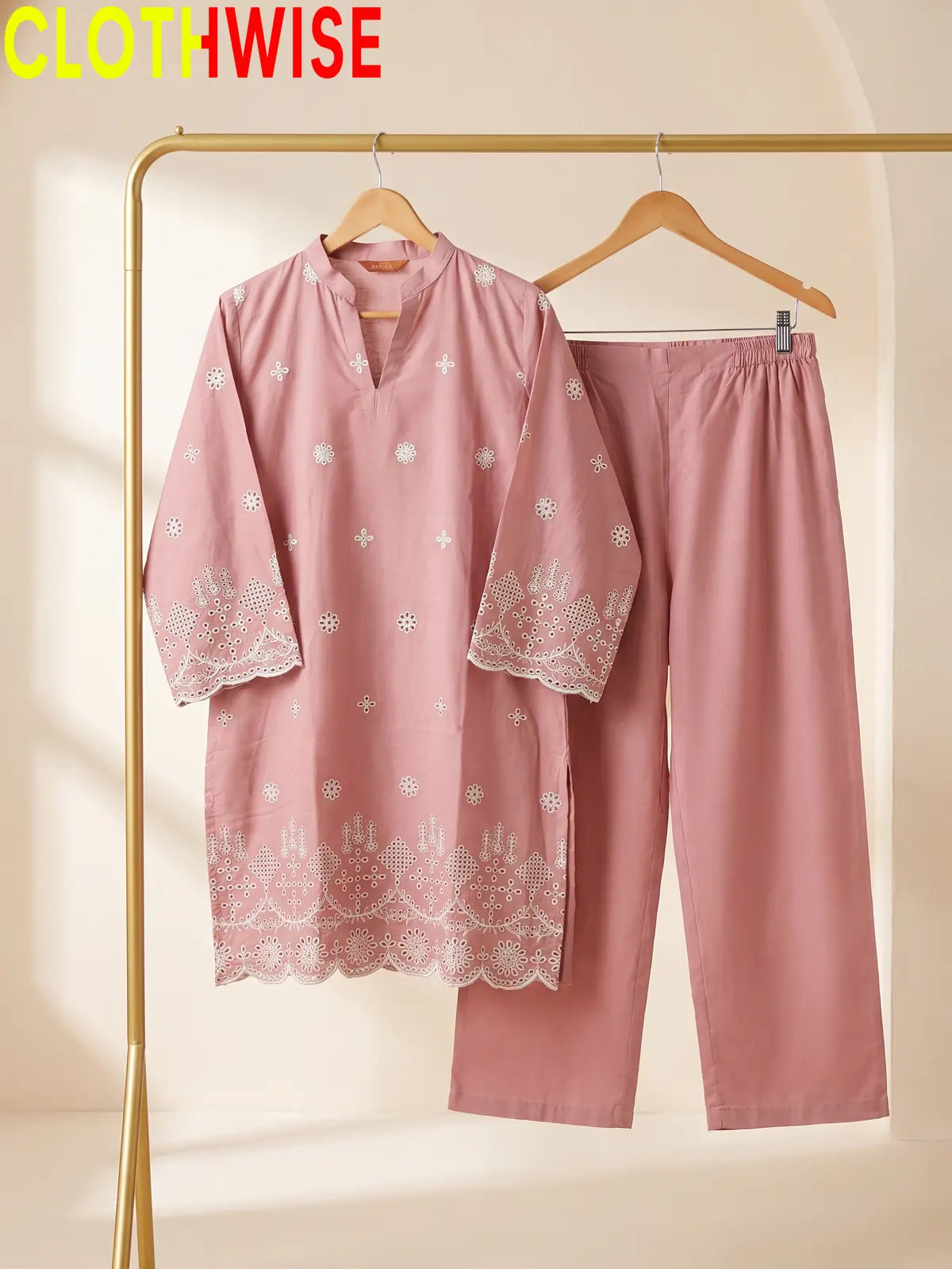 A two-piece women's pajama set in dusty pink, featuring a long-sleeve tunic with white floral embroidery and lace trim, paired with matching straight-leg pants. The set is displayed on wooden hangers hanging from a gold clothing rack against a warm beige background.