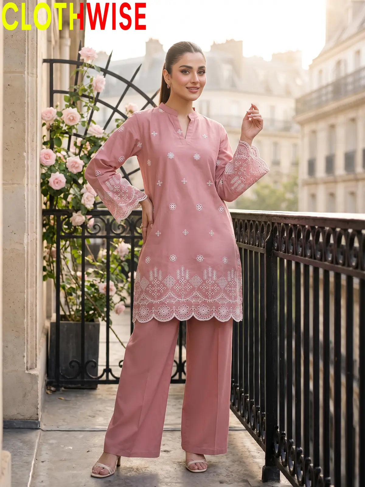 A woman modeling a dusty pink two-piece kurta set with white embroidery and lace hem, standing on a balcony with a black railing and pink roses in the background. The brand name CLOTHWISE is displayed in the top left corner.