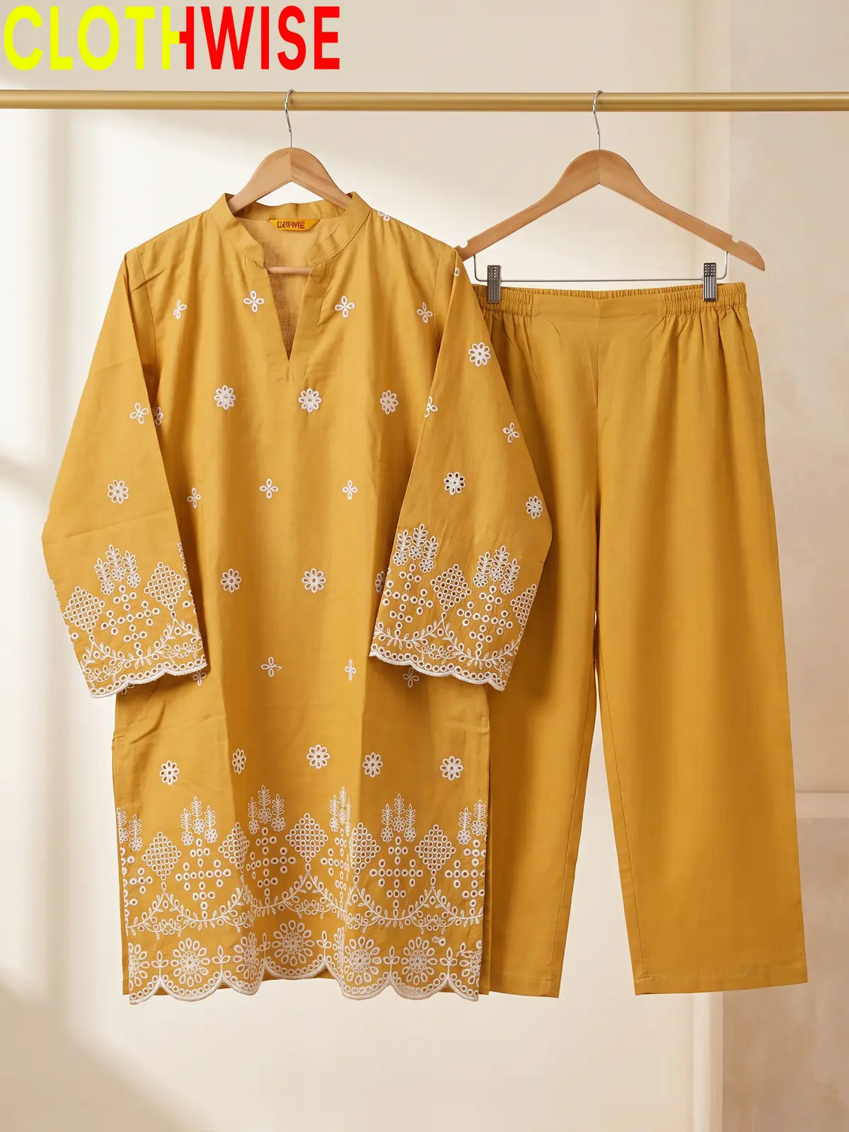 A mustard yellow two-piece clothing set from the brand CLOTHWISE, featuring a long-sleeve tunic with white embroidered patterns and matching straight-leg pants, displayed on wooden hangers.