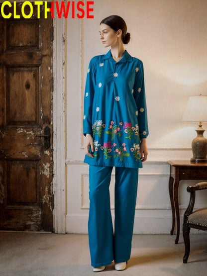 Woman wearing a teal outfit with floral patterns in a room with wooden door and lamp.