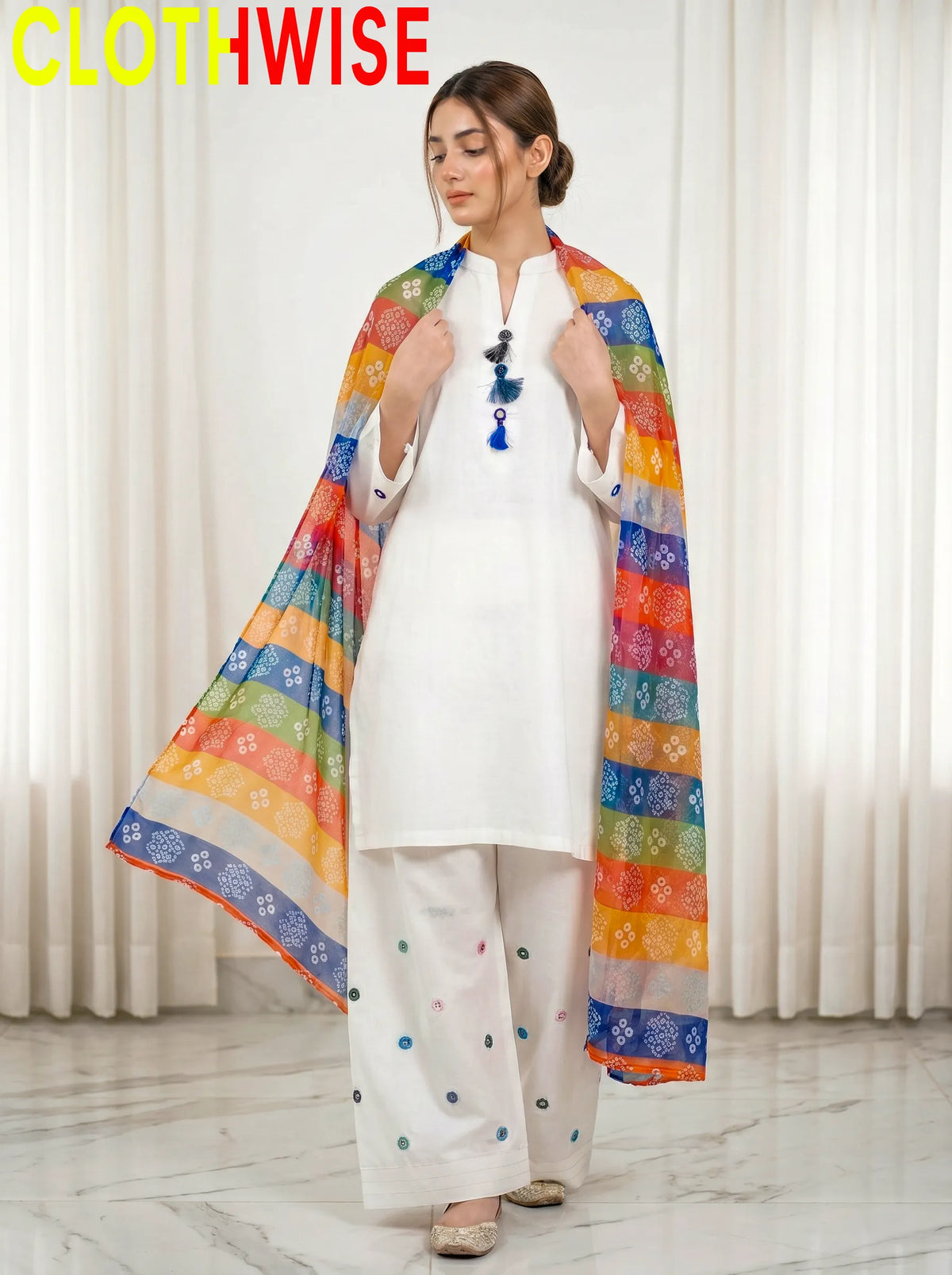 Woman wearing a white outfit with a colorful scarf, standing against a white curtain background.